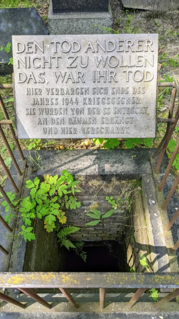 Steintafel mit der Inschrift Den Tod anderer nicht zu wollen über einem gemauerten Schacht, eingefasst von einem rostigen Eisengeländer