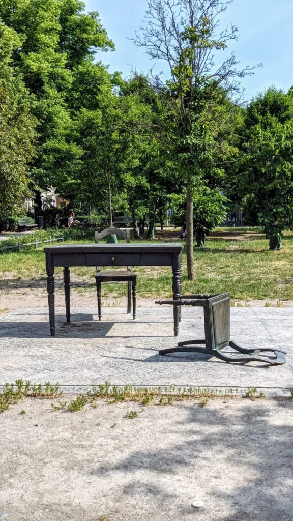 Bronzedenkmal Der verlassene Raum auf dem Koppenplatz: ein Tisch, ein Stuhl und ein umgestürzter Stuhl auf einer Bodenplatte im Gras