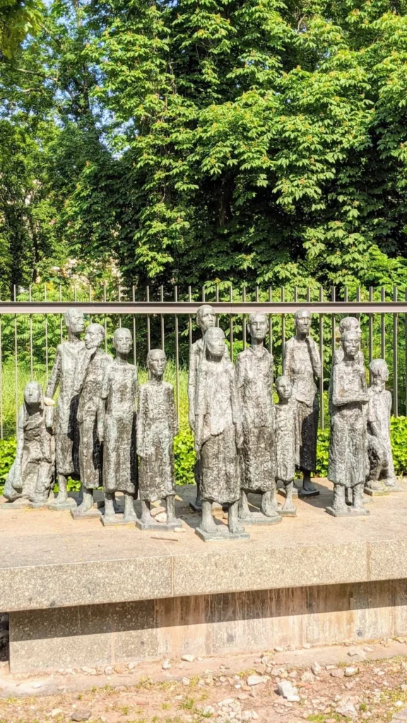 Bronzene Figurengruppe aus dreizehn schmalen Gestalten auf einem Steinsockel vor Kastanien