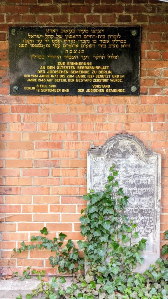 Schwarze Gedenktafel von 1948 an einer Backsteinmauer, darunter ein alter, von Efeu überwachsener Grabstein