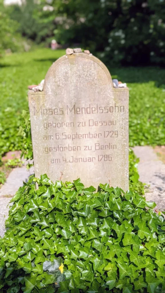 Heller Grabstein mit Rundbogen für Moses Mendelssohn, darauf kleine abgelegte Steine, davor Efeu