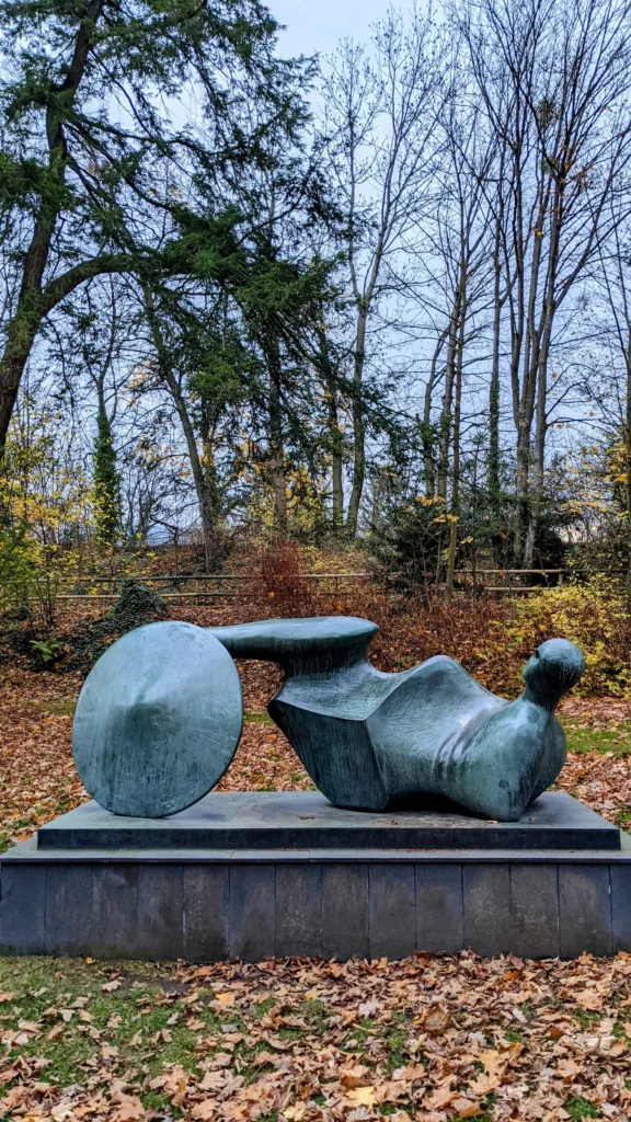Liegende Bronzefigur mit großer Schildscheibe auf niedrigem Sockel zwischen Herbstlaub und Bäumen