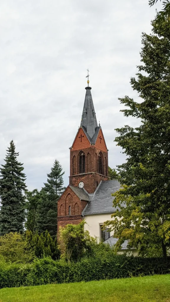 Jesuskirche am Anger in Kaulsdorf mit rotem Backsteinturm, hellem Kirchenschiff und Nadelbäumen davor