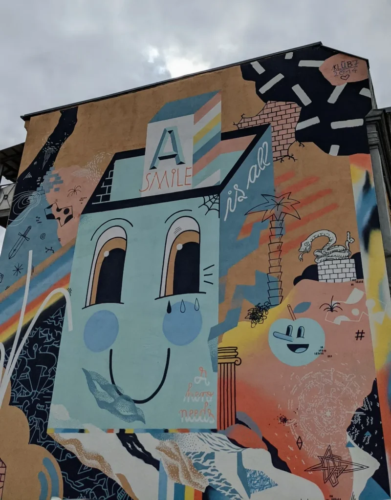 Wandbild von Klub7 an der Berliner Straße in Lutherstadt Wittenberg: Haus mit Gesicht und verspielte Figuren in Blau, Rosa und Orange auf einer Giebelwand