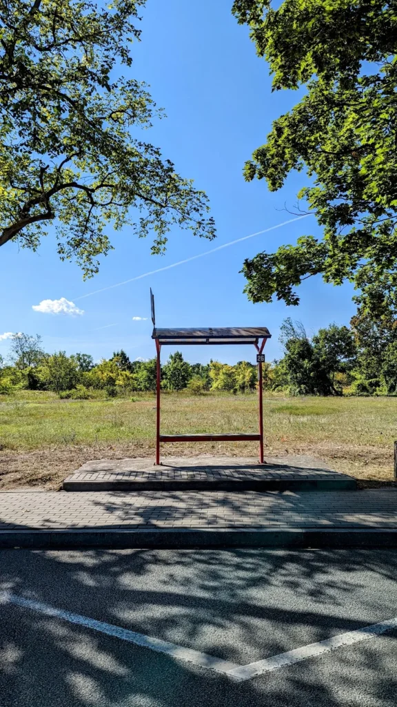 Rotes Metallgestell mit schmalem Dach und der Nummer 26 auf einem gepflasterten Podest an einem Parkplatz in Kostrzyn nad Odrą, dahinter eine Wiese