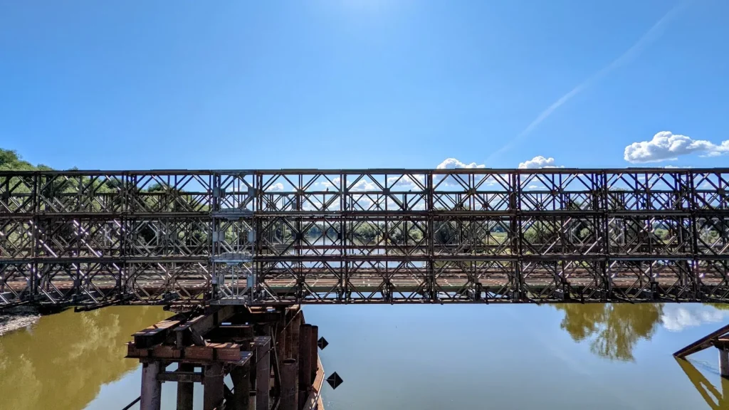 Rostbraunes Stahlfachwerk dicht über dem braunen Wasser der Warthe in Kostrzyn nad Odrą, davor ein alter Pfeilerrest