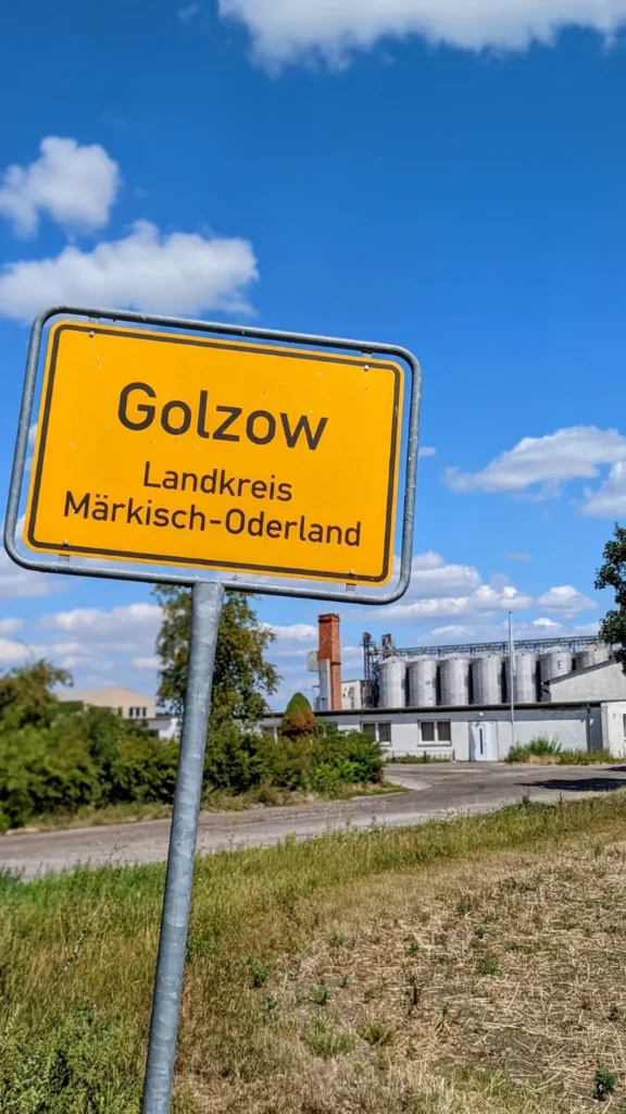Gelbes Ortsschild Golzow, Landkreis Märkisch-Oderland, dahinter silberne Getreidesilos und ein Backsteinschornstein