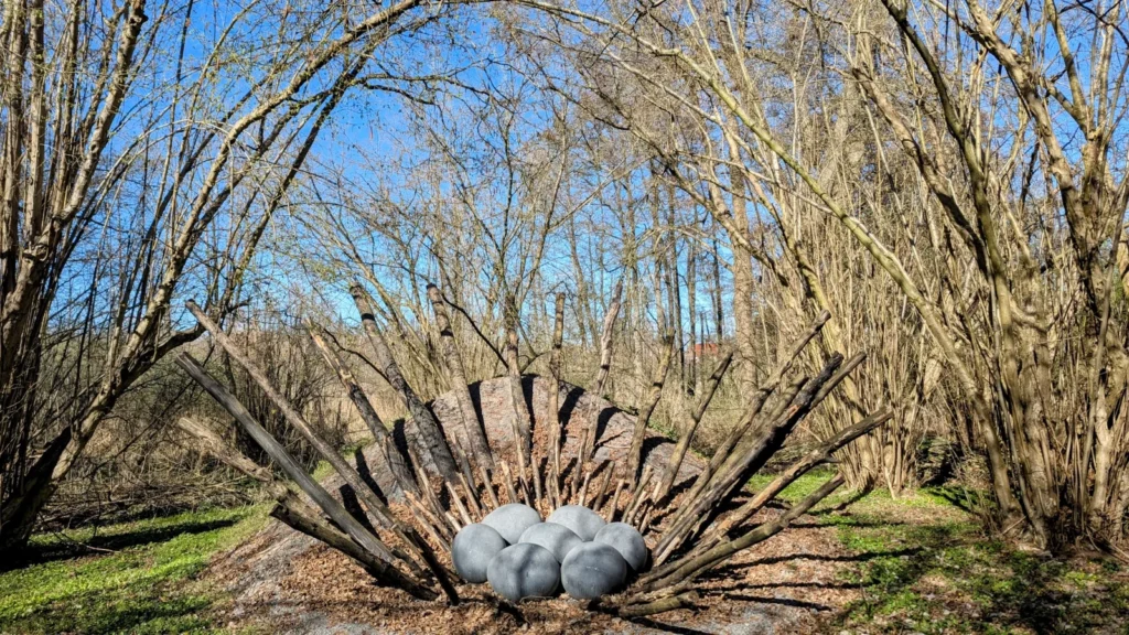 Skulptur in Form eines Nests aus Holzbalken mit grauen Eiern zwischen Sträuchern in Schwante