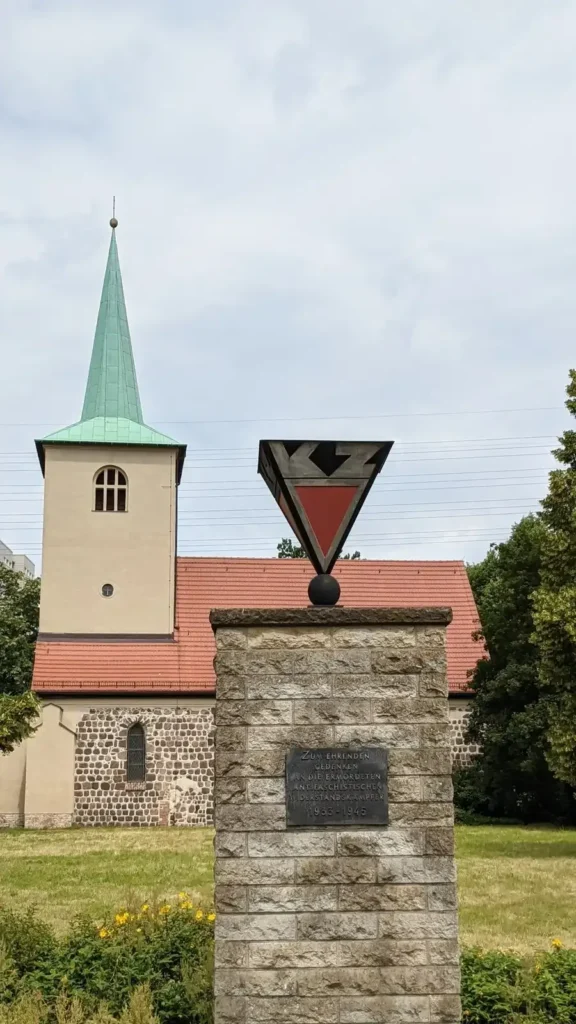 Feldsteinkirche mit Turm am Loeperplatz in Lichtenberg, davor ein Gedenkstein mit rotem Dreieck