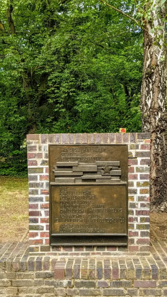 Backsteinsockel mit Bronzetafel zwischen Bäumen im hinteren Teil des Zentralfriedhofs Friedrichsfelde