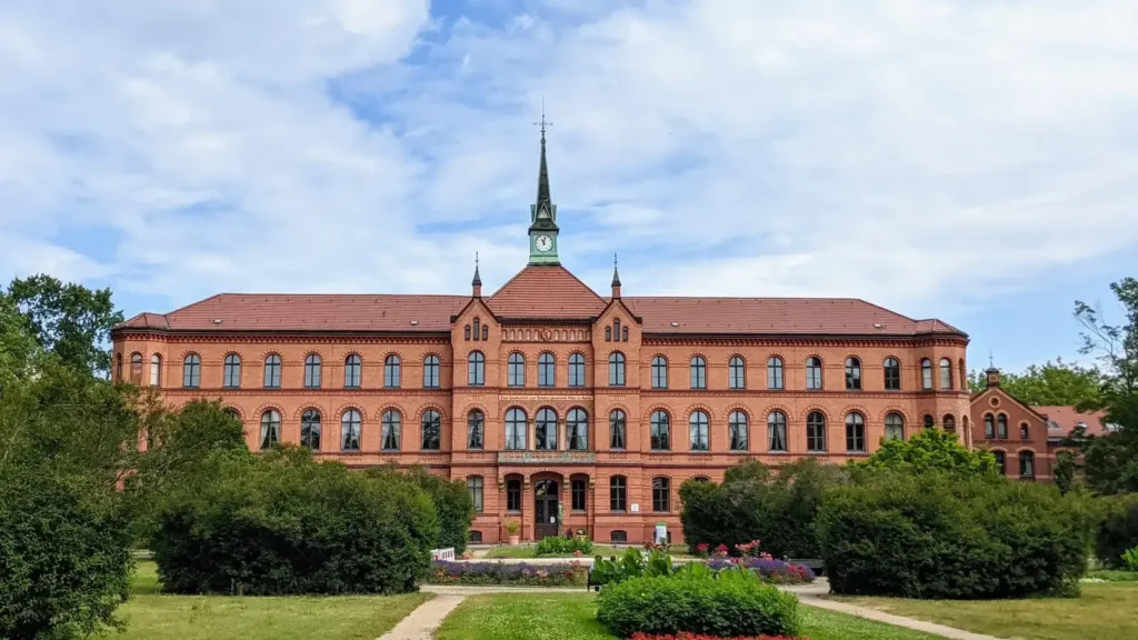 Großes rotes Backsteingebäude mit Mittelturm des Krankenhauses Herzberge hinter Rasen und Beeten