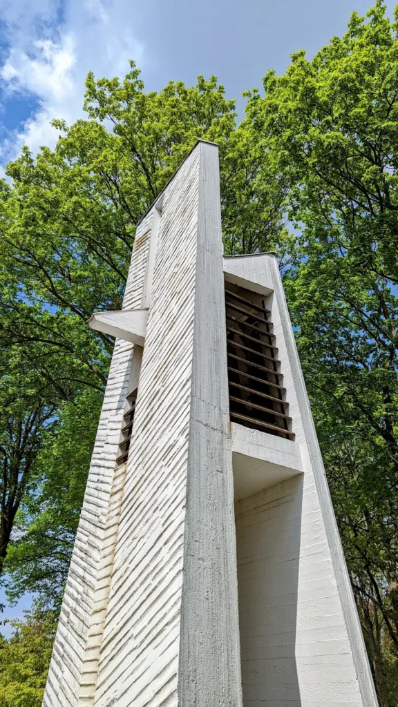 Weißer schlanker Turm aus Sichtbeton mit Schallöffnung zwischen hohen Laubbäumen, Lichtenrade