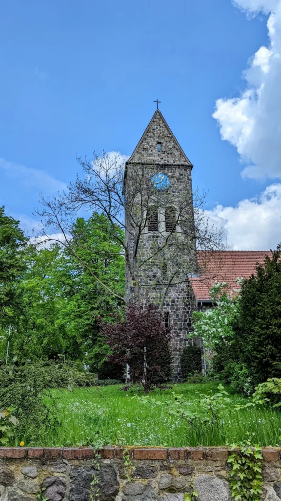 Feldsteinturm der Dorfkirche Lichtenrade mit Uhr und Satteldach, davor Kirchhofmauer und Wiese