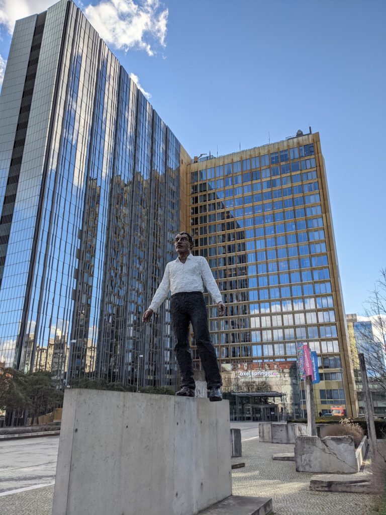 Skulptur eines Mannes, der auf einem Mauerstück balanciert, dahinter die Glasfassaden des Axel-Springer-Hochhauses