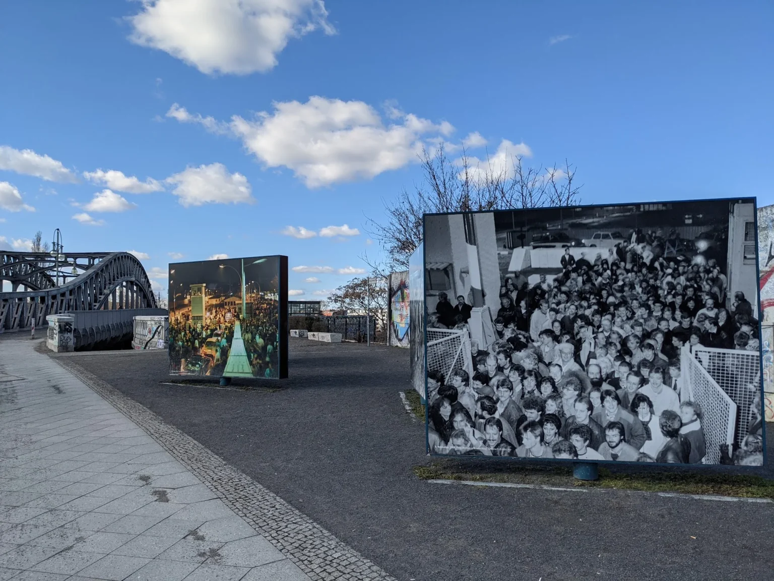 Großformatige Fototafeln zum Mauerfall auf einem gepflasterten Platz, im Hintergrund die Stahlbögen der Bösebrücke