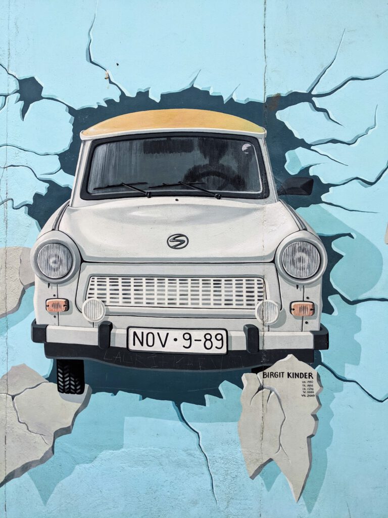 Wandbild an der East Side Gallery: Ein weißer Trabant durchbricht die Mauer, auf dem Kennzeichen steht „NOV 9-89“