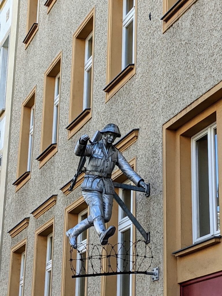 Metallskulptur eines springenden Grenzsoldaten über Stacheldraht an einer Hausfassade