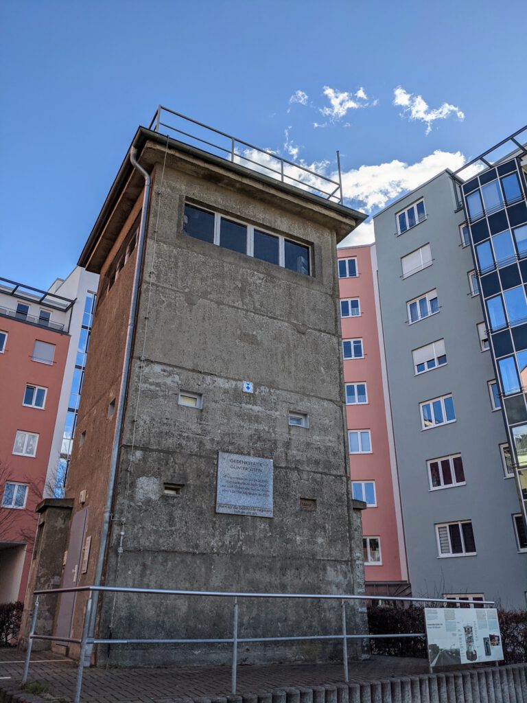Grauer Wachturm aus Beton mit Fensterband steht zwischen bunten Wohnhäusern