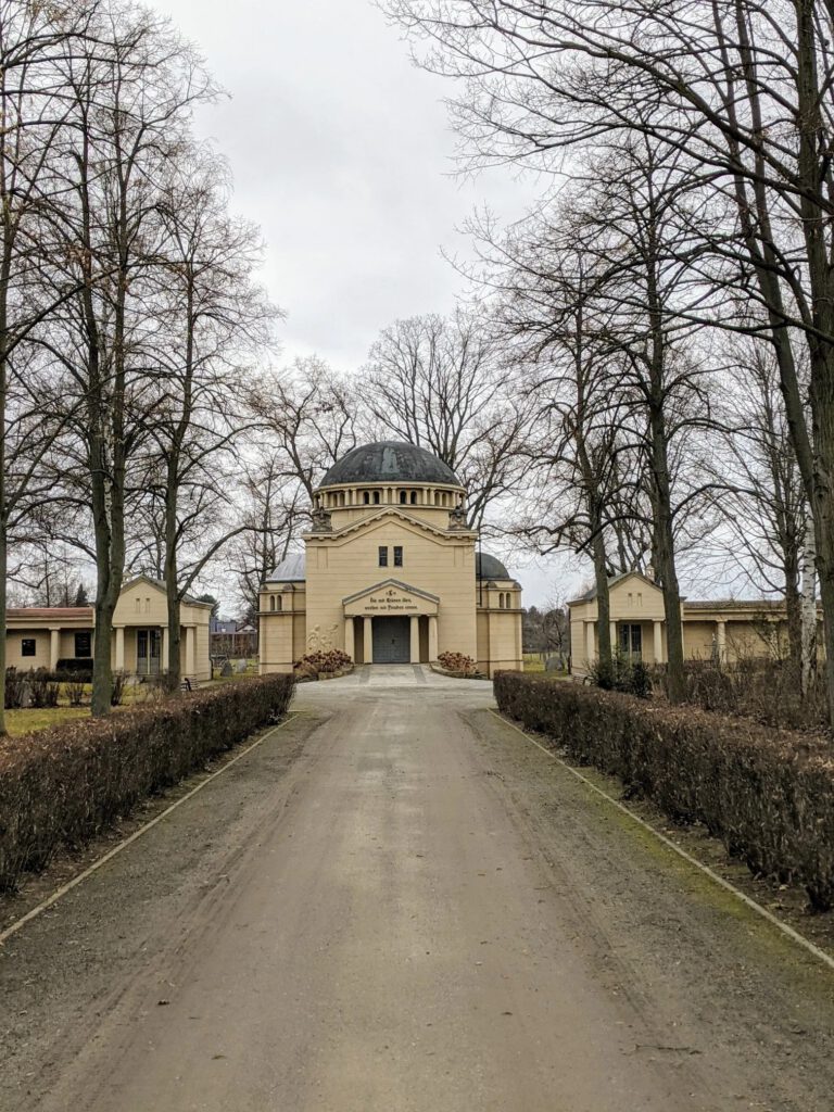 Gelbe Feierhalle mit Kuppel am Ende eines von Hecken und kahlen Bäumen gesäumten Wegs