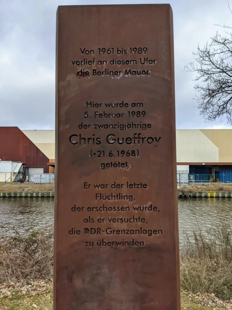 Rostfarbene Stahlstele mit Inschrift für Chris Gueffroy am Ufer eines Kanals, dahinter Fabrikhallen