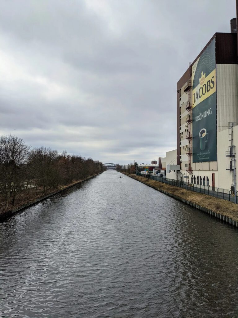 Blick von einer Brücke über einen schnurgeraden Kanal, rechts ein Fabrikgebäude mit großem Werbebanner