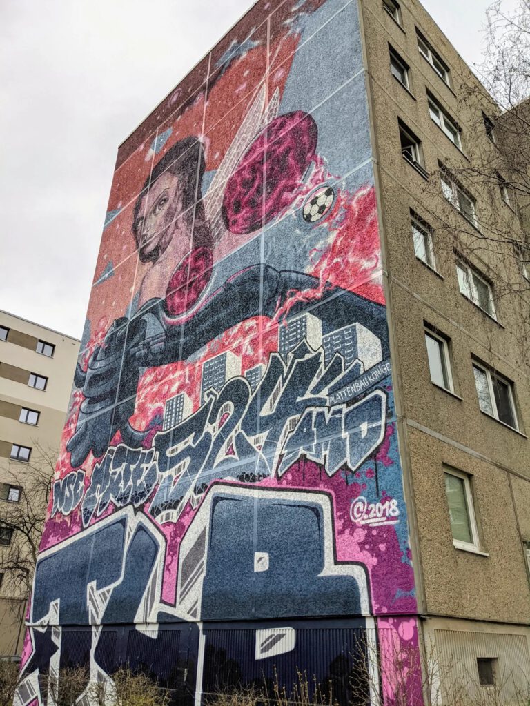 Buntes Wandbild über die ganze Giebelseite eines Plattenbaus mit einer Frau, Planeten, einem Fußball und Graffiti-Schriftzügen