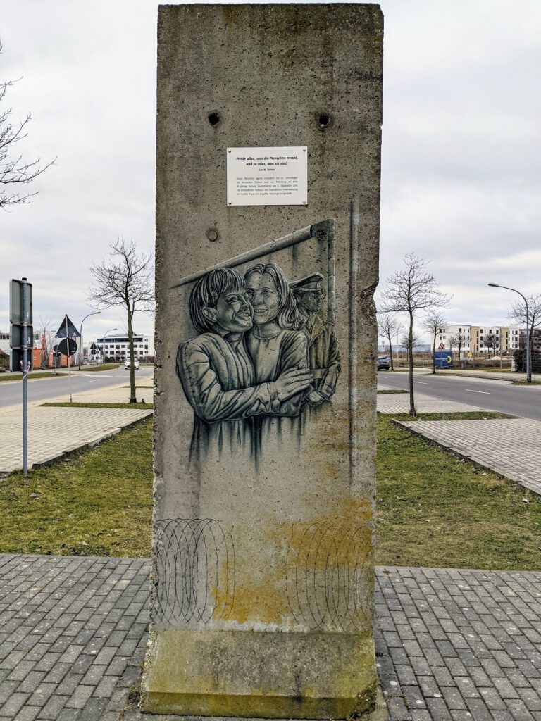 Einzelnes Mauersegment mit einem gemalten Bild zweier Menschen, die sich umarmen, darüber eine kleine Tafel, dahinter Straßen und Neubauten