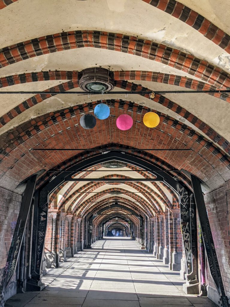 Arkadengang der Oberbaumbrücke mit Backsteinbögen, unter dem Gewölbe hängen vier farbige Scheiben