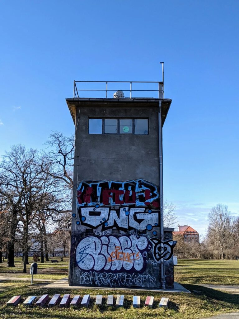 Grauer Wachturm mit Graffiti am Sockel steht auf einer Wiese unter blauem Himmel