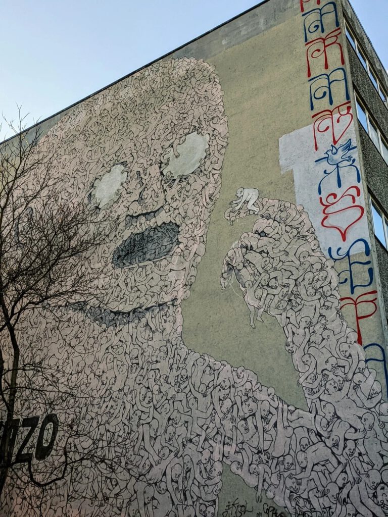 Riesiges Wandbild an einer Brandwand: eine Figur, die aus vielen kleinen nackten Menschen zusammengesetzt ist und einen einzelnen Menschen in der Hand hält