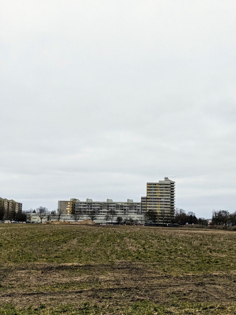Wohnhochhäuser am Stadtrand hinter einem weiten, kahlen Feld unter grauem Himmel