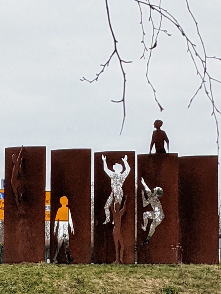 Fünf rostige Stahlstelen, an denen ausgeschnittene Figuren hochklettern, eine sitzt oben auf