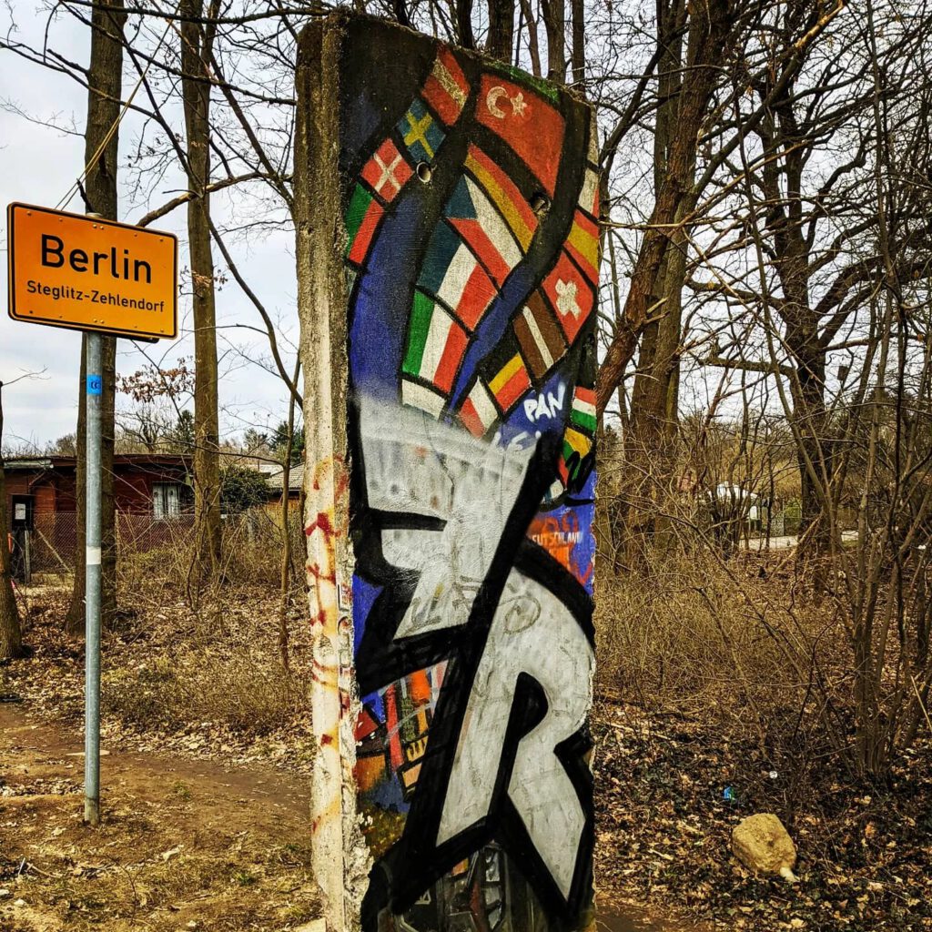 Bemaltes Mauersegment mit Flaggen und Graffiti neben dem Ortsschild „Berlin Steglitz-Zehlendorf“