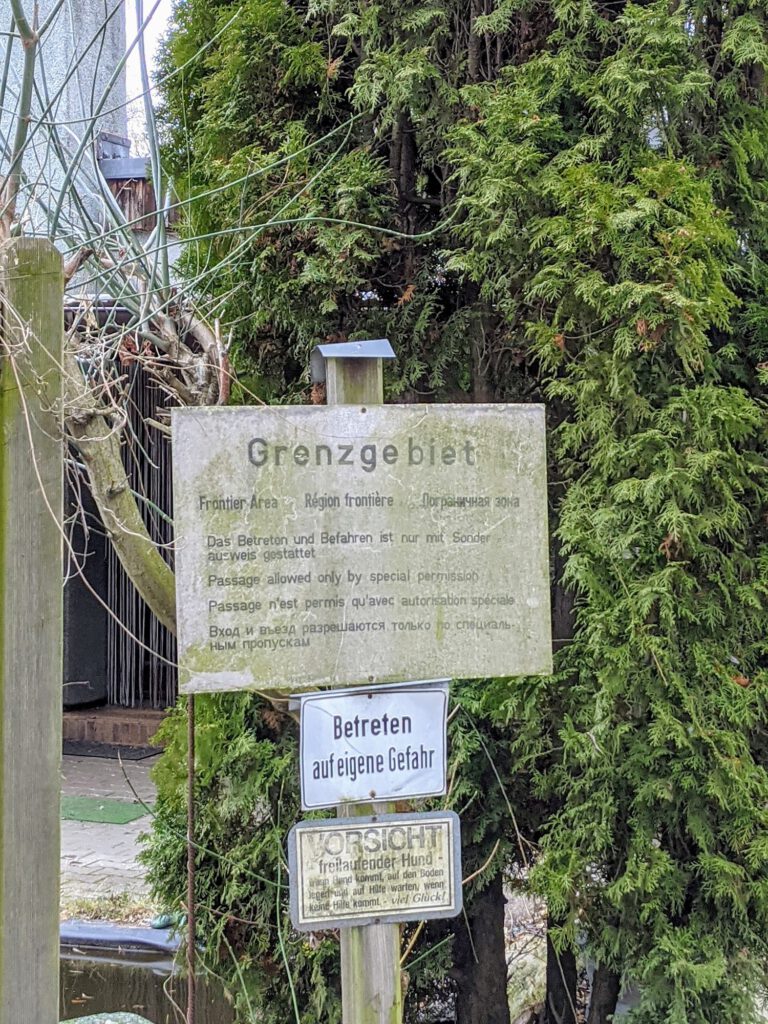 Verwittertes Schild mit der Aufschrift „Grenzgebiet“ vor einer Hecke, darunter zwei kleinere Warnschilder