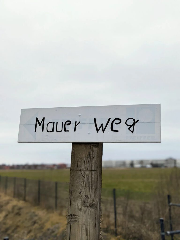 Weißes Schild mit der handgeschriebenen Aufschrift „Mauer Weg“ auf einem Holzpfosten vor einem Feld