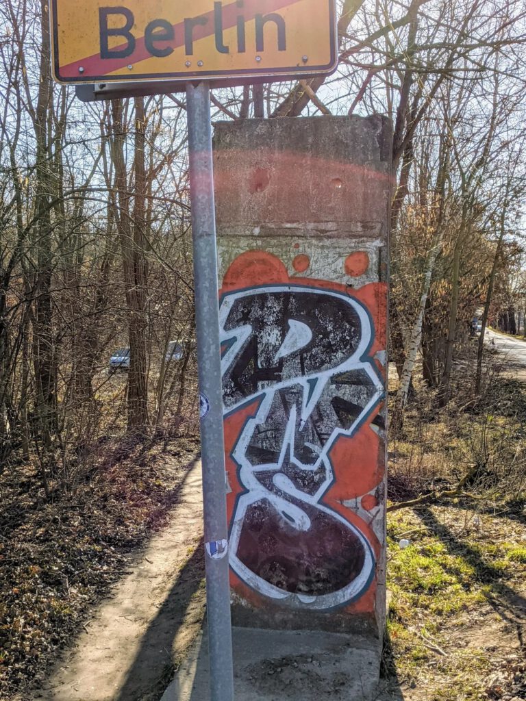 Mauersegment mit Graffiti hinter einem durchgestrichenen Ortsschild „Berlin“ an einem Pfad