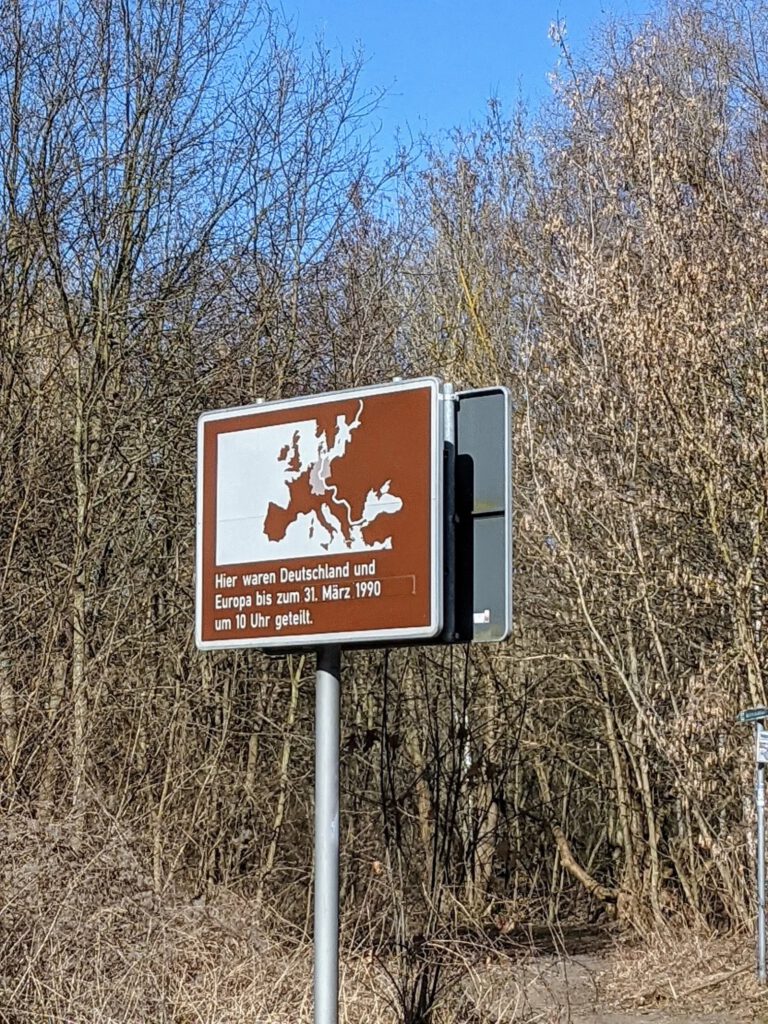 Zwei bemalte Mauersegmente im Gegenlicht auf einer Wiese mit kahlen Bäumen, eines zeigt eine Kirche und einen Wachturm