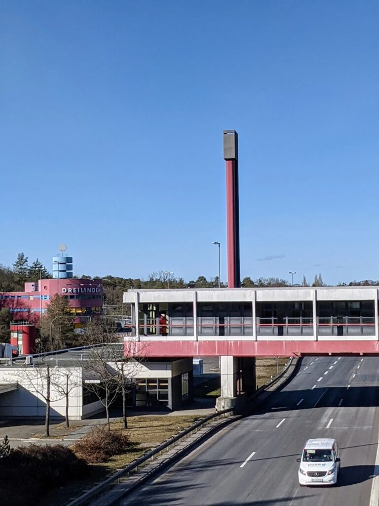 Gelbes Ortsschild: oben „Berlin“ mit Pfeil, unten durchgestrichen „Teltow“, im Hintergrund Wasser