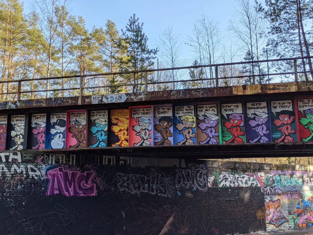 Rostige Stahlbrücke im Kiefernwald, deren Träger mit einer Reihe bunter Comicfiguren mit Sprechblasen bemalt ist, darunter besprühte Backsteinmauern