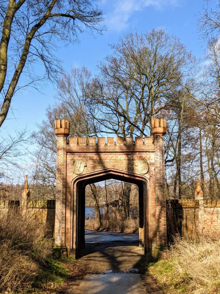 Tor aus gelbrotem Backstein mit Zinnen, zwei Türmchen und Adlerwappen, dahinter ein Weg zum Wasser