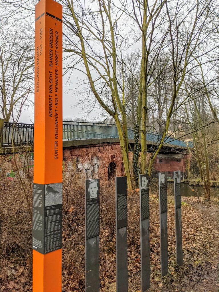 Orangefarbene Stele mit fünf Namen, daneben fünf schmale Tafeln mit Porträtfotos, dahinter eine Brücke aus rotem Stein