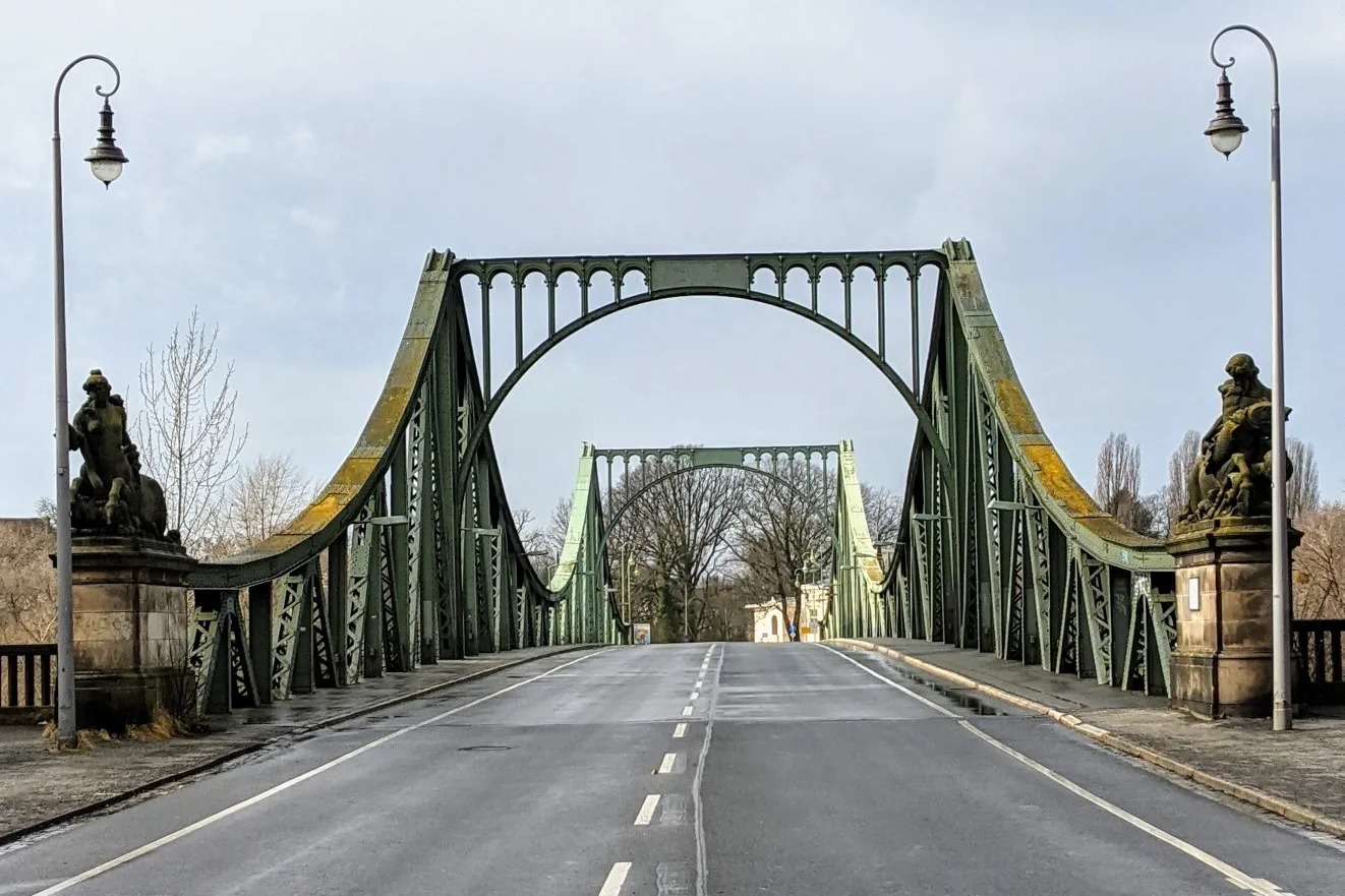 Blick über die leere Fahrbahn auf die grünen Stahlbögen der Glienicker Brücke, links und rechts Laternen und Sandsteinfiguren
