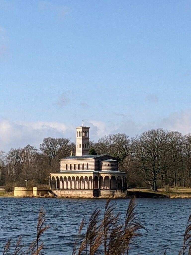 Kirche mit Arkadengang und freistehendem Glockenturm direkt am gegenüberliegenden Ufer, im Vordergrund Wasser und Schilf