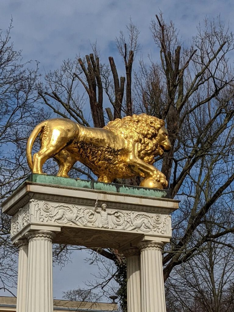 Vergoldeter Löwe mit der Pranke auf einer Kugel steht auf einem weißen Säulenpodest vor blauem Himmel