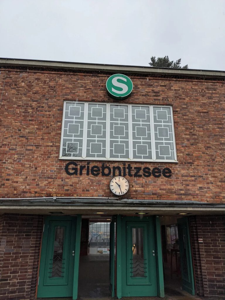 Backsteinfassade des Bahnhofs mit grünem S-Bahn-Zeichen, Schriftzug „Griebnitzsee“, Uhr und grünen Türen