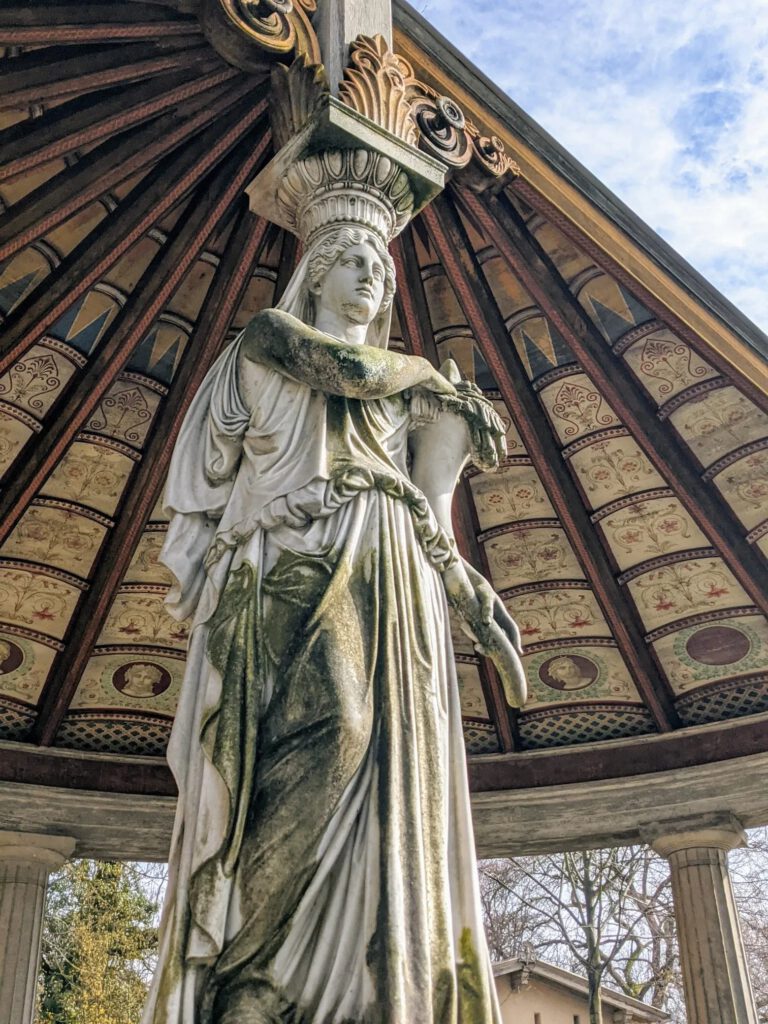 Weiße, bemooste Frauenfigur trägt als Stütze ein rundes, innen bunt bemaltes Holzdach