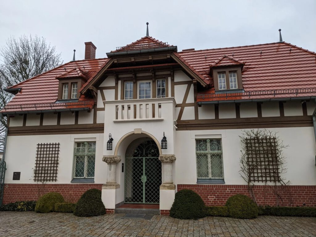 Weiß verputzte Villa mit braunem Fachwerk, rotem Ziegeldach und einem Rundbogenportal mit grünem Gitter