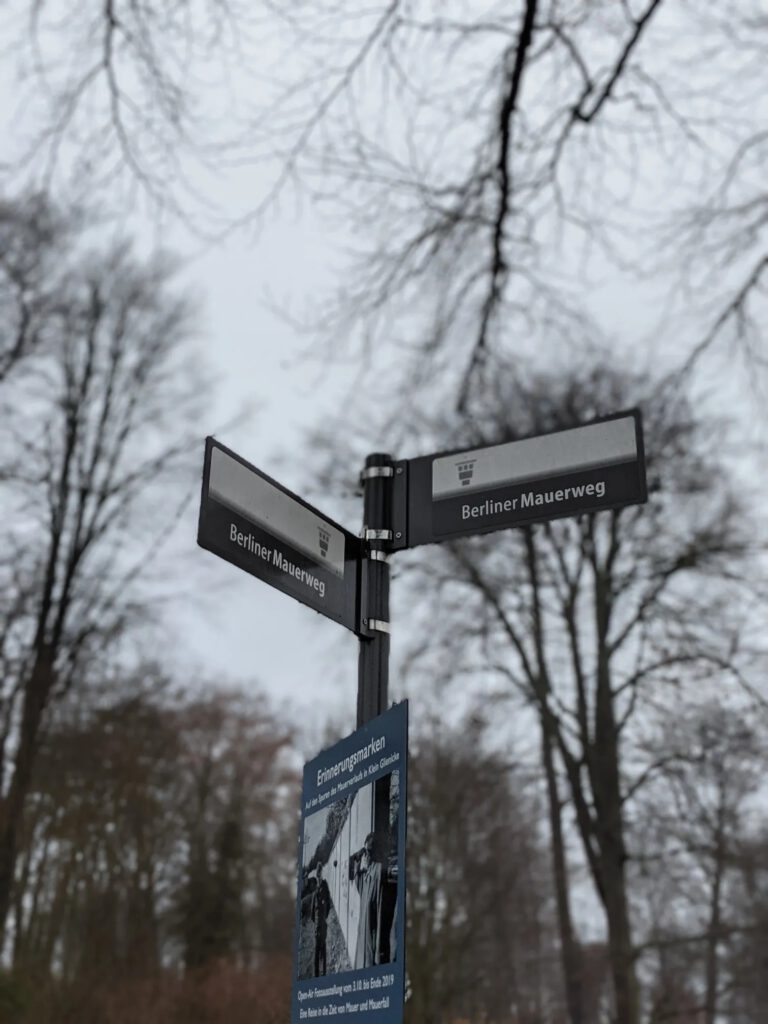Zwei graue Wegweiser mit der Aufschrift „Berliner Mauerweg“ vor kahlen Bäumen, darunter ein blaues Schild mit dem Titel „Erinnerungsmarken“