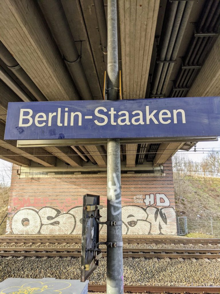 Blaues Bahnhofsschild „Berlin-Staaken“ unter einer Betonbrücke, dahinter Gleise und ein Graffiti