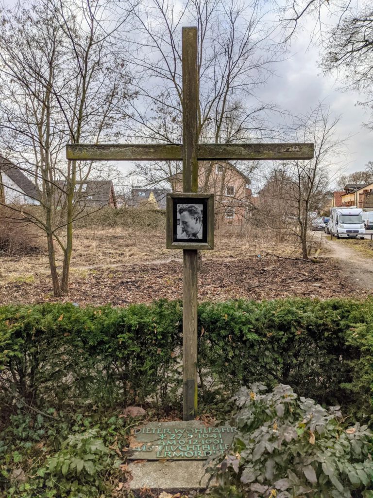 Holzkreuz mit einem gerahmten Porträtfoto, davor eine Bodenplatte mit dem Namen Dieter Wohlfahrt und seinen Lebensdaten